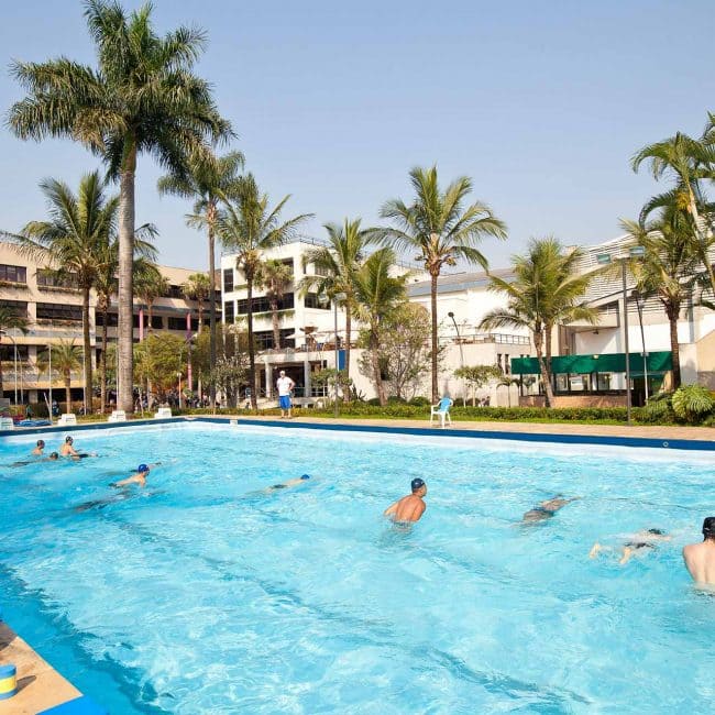 Vista de uma piscina com várias pessoas nadando sob palmeiras em um dia ensolarado, com um edifício ao fundo.