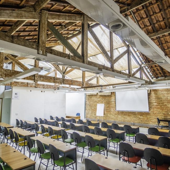 Sala de aula com mesas e cadeiras organizadas, teto de madeira exposta e paredes de tijolo à vista.